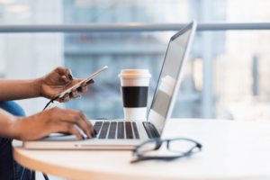 Foto van een persoon achter laptop met mobiele telefoon in de hand, bekertje koffie staat naast laptop