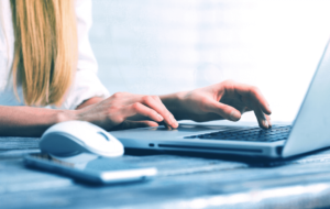 De afbeelding toont een persoon die typt op een laptop, met een witte computermuis en een smartphone op een houten bureau in de buurt.