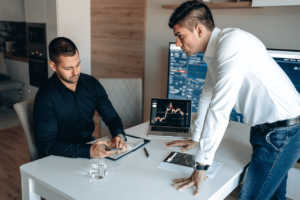 Foto van 2 personen die overleggen bij een bureau met laptop en groot scherm met statistieken